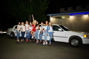 Cool bride in red dress posing with her bridesmaids against the limousine at bachelorette party in San Diego, CA
